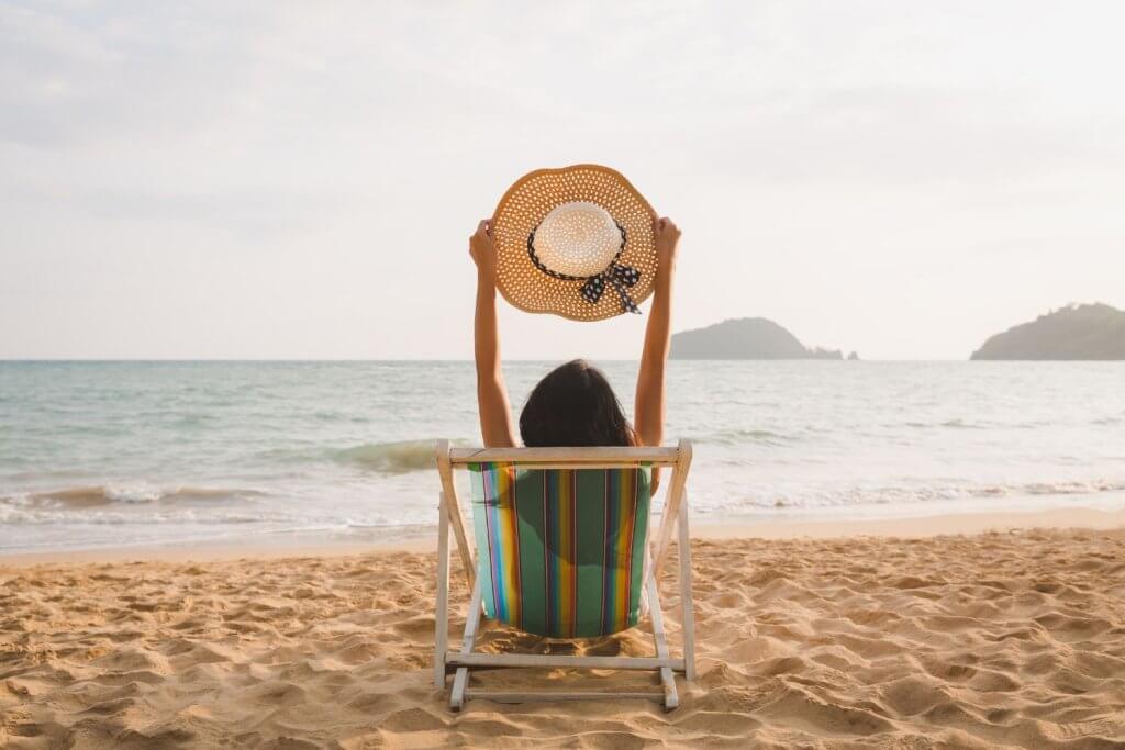 Femme assise face à la mer sur une plage, dans un fauteuil en bois, au coucher du soleil.